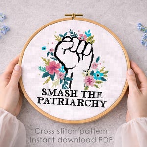May include: A circular wooden embroidery hoop containing a white fabric with a cross-stitch design. The design features a black outlined raised fist surrounded by pink and blue flowers and green leaves, with the text SMASH THE PATRIARCHY below.