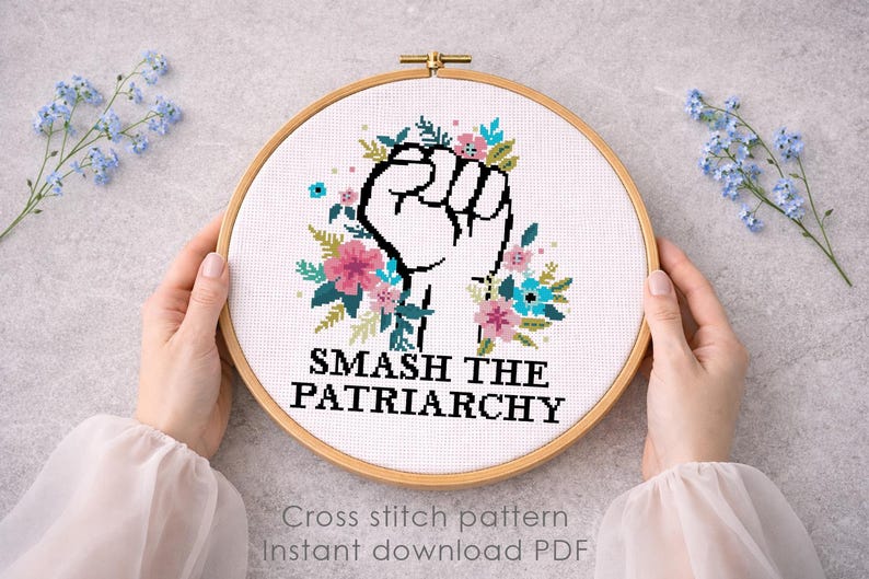 May include: A circular wooden embroidery hoop containing a white fabric with a cross-stitch design. The design features a black outlined raised fist surrounded by pink and blue flowers and green leaves, with the text SMASH THE PATRIARCHY below.