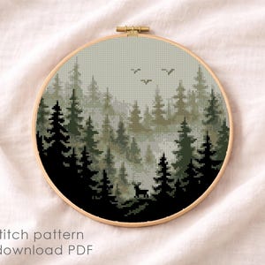 May include: A cross-stitch pattern of a forest scene in a round wooden hoop. The design features dark green and black trees, a deer, and birds against a misty, light green background. The text "Cross stitch pattern Instant download PDF" is at the bottom.