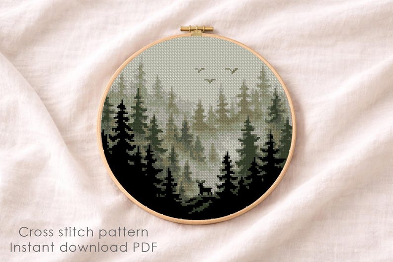 May include: A cross-stitch pattern of a forest scene in a round wooden hoop. The design features dark green and black trees, a deer, and birds against a misty, light green background. The text "Cross stitch pattern Instant download PDF" is at the bottom.