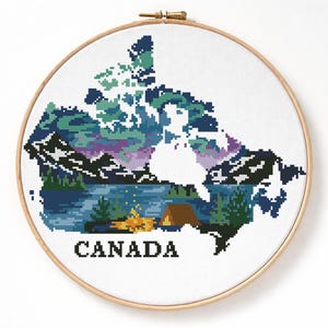 May include: Cross-stitch embroidery of Canada's map, featuring a landscape with mountains, a lake, and a campfire. The design uses shades of blue, green, and purple. The word "CANADA" is stitched at the bottom. The embroidery is set in a wooden hoop.