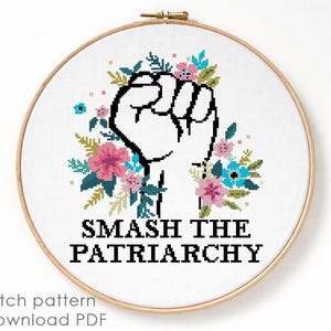 May include: A circular wooden embroidery hoop containing a white fabric with a cross-stitch design. The design features a black outlined raised fist surrounded by pink and blue flowers and green leaves, with the text SMASH THE PATRIARCHY below.