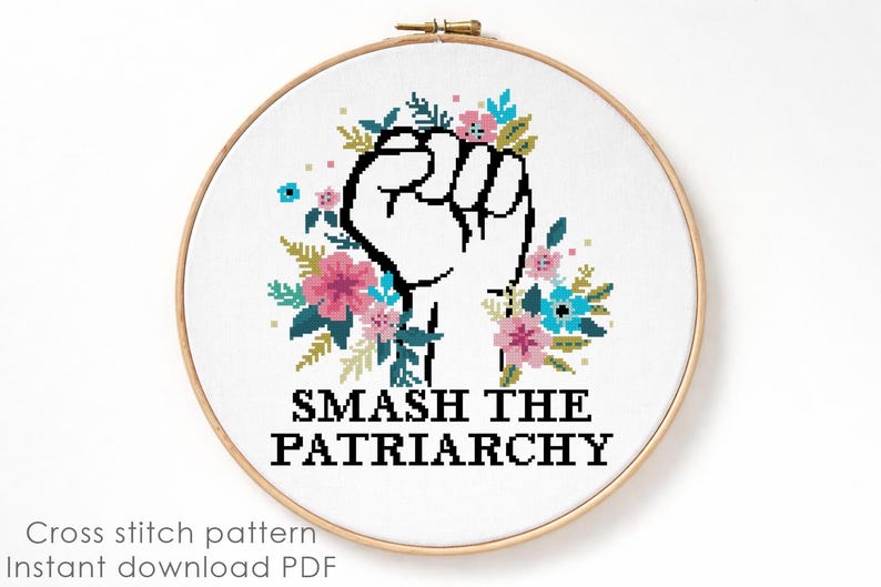 May include: A circular wooden embroidery hoop containing a white fabric with a cross-stitch design. The design features a black outlined raised fist surrounded by pink and blue flowers and green leaves, with the text SMASH THE PATRIARCHY below.