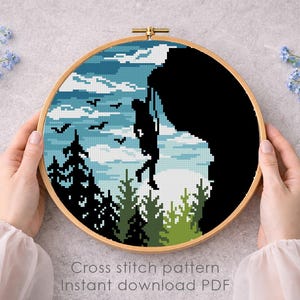 Op de afbeelding: Een kruissteekpatroon in een houten hoepel toont een bergbeklimmer op een klif. Het ontwerp toont een blauwe lucht met vogels, bomen en de tekst "Cross stitch pattern Instant download PDF".