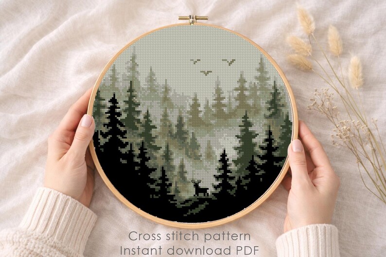 May include: A cross-stitch pattern of a forest scene in a wooden hoop. The design features dark green and black trees, a deer, and birds against a misty background. The text "Cross stitch pattern Instant download PDF" is visible.