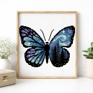 Puede incluir: Arte de punto de cruz de una mariposa con un dise&ntilde;o de cielo nocturno. La mariposa tiene alas azules y moradas con un patr&oacute;n de cielo estrellado. El centro de la mariposa presenta una silueta de bosque. La obra de arte est&aacute; enmarcada en un marco de madera.