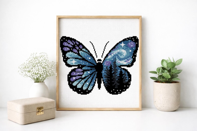 Puede incluir: Arte de punto de cruz de una mariposa con un dise&ntilde;o de cielo nocturno. La mariposa tiene alas azules y moradas con un patr&oacute;n de cielo estrellado. El centro de la mariposa presenta una silueta de bosque. La obra de arte est&aacute; enmarcada en un marco de madera.