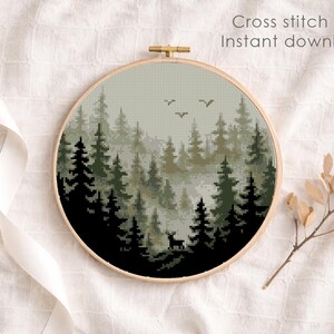 May include: A round embroidery hoop featuring a cross-stitch pattern of a forest scene with trees, a deer, and birds. The text "Cross stitch pattern Instant download PDF" is visible. The hoop is set against a white fabric background.