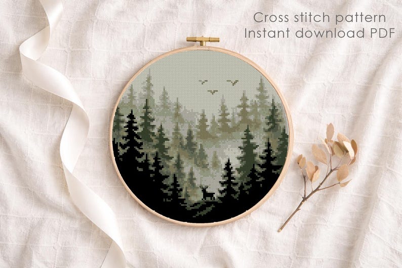 May include: A round embroidery hoop featuring a cross-stitch pattern of a forest scene with trees, a deer, and birds. The text "Cross stitch pattern Instant download PDF" is visible. The hoop is set against a white fabric background.
