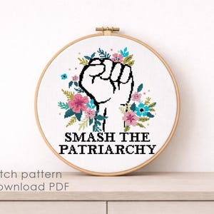 May include: A circular wooden embroidery hoop containing a cross-stitch design on white fabric. The design features a black outlined raised fist surrounded by pink, blue, and green floral elements, with the text SMASH THE PATRIARCHY in black.