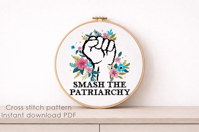 May include: A circular wooden embroidery hoop containing a cross-stitch design on white fabric. The design features a black outlined raised fist surrounded by pink, blue, and green floral elements, with the text SMASH THE PATRIARCHY in black.