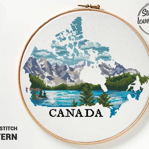 Peut inclure: Un motif de point de croix du Canada, représentant un paysage de montagnes, un lac et des arbres. Le mot "CANADA" est brodé en lettres noires. Le motif est encadré dans un cercle à broder en bois. Le texte "CROSS STITCH PATTERN" est en dessous.
