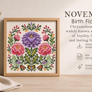 May include: A framed cross-stitch pattern featuring a floral design with a central purple flower and two pink flowers. The text "NOVEMBER Birth Flowers" is at the top, with additional text below. The design includes green leaves and birds.