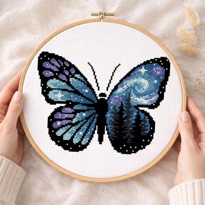 Puede incluir: Bordado de punto de cruz de una mariposa con un dise&ntilde;o de cielo nocturno. La mariposa tiene alas azules y moradas con un patr&oacute;n de estrellas y una silueta de bosque. El bordado est&aacute; en un bastidor de madera.