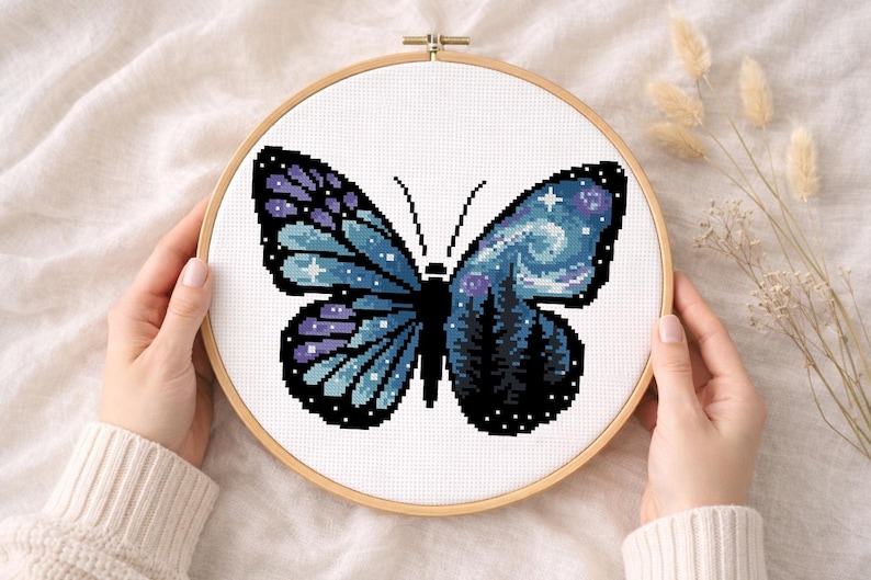 Puede incluir: Bordado de punto de cruz de una mariposa con un dise&ntilde;o de cielo nocturno. La mariposa tiene alas azules y moradas con un patr&oacute;n de estrellas y una silueta de bosque. El bordado est&aacute; en un bastidor de madera.
