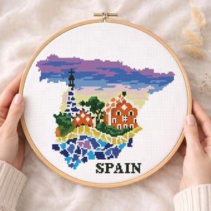 Puede incluir: Un bastidor de bordado de punto de cruz con un mapa colorido de Espa&ntilde;a. El dise&ntilde;o incluye monumentos y la palabra "SPAIN" en negro. El bordado est&aacute; en un bastidor de madera, sostenido por manos.