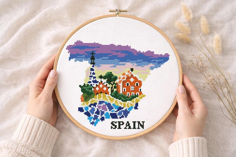 Puede incluir: Un bastidor de bordado de punto de cruz con un mapa colorido de Espa&ntilde;a. El dise&ntilde;o incluye monumentos y la palabra "SPAIN" en negro. El bordado est&aacute; en un bastidor de madera, sostenido por manos.