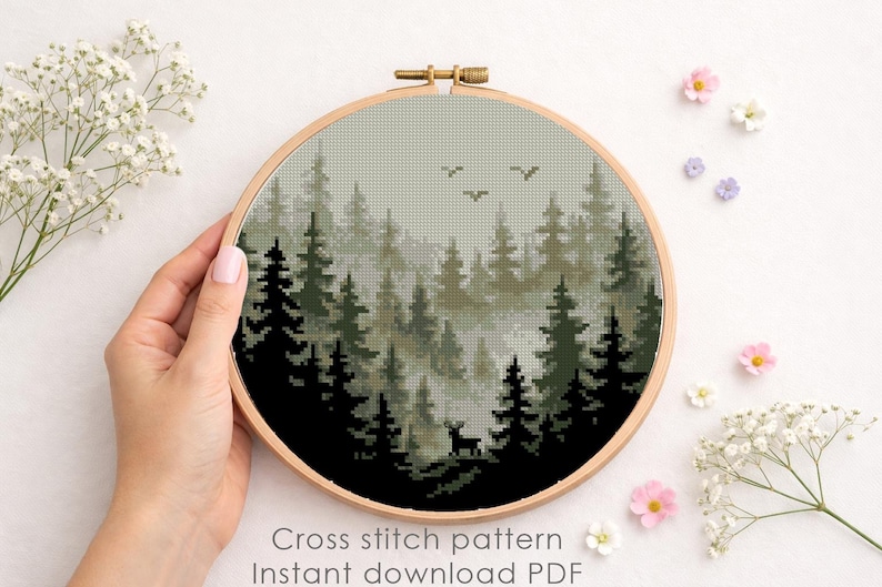 May include: A cross-stitch pattern of a forest scene in a wooden embroidery hoop. The design features dark green trees, a deer, and birds against a misty background. The text "Cross stitch pattern Instant download PDF" is visible.