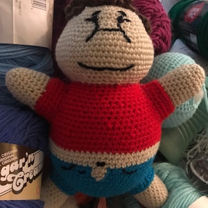 Puede incluir: Una muñeca de ganchillo hecha a mano con cabeza beige, cabello castaño, camisa roja y pantalones cortos azules. La muñeca tiene ojos cosidos negros y una boca sonriente. Está rodeada de ovillos de lana de varios colores.