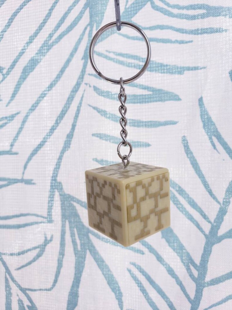 May include: A beige cube keychain with a geometric pattern. The keychain is attached to a silver chain and a silver ring.