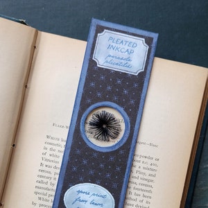 Puede incluir: Un marcapáginas hecho a mano con un diseño de hongo Pleated Inkcap. El marcapáginas tiene un fondo azul oscuro con un patrón repetido y dos óvalos azul claro con texto. Un recorte circular revela una impresión de esporas.
