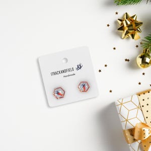Puede incluir: Un par de pendientes de botón hexagonales con fondo blanco y un diseño rojo y azul en el interior. Los pendientes están en una tarjeta blanca con el texto "ITRACKANDFIELD Handmade".