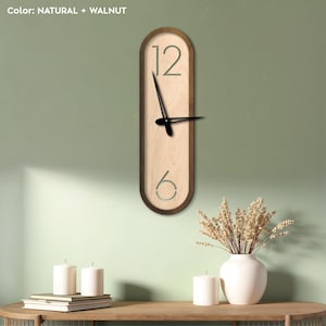 Puede incluir: Un reloj de pared de madera con un acabado de madera natural y un marco de nogal. La esfera del reloj es de forma ovalada y presenta un diseño sencillo con agujas negras y números blancos. El reloj está colgado en una pared verde claro.