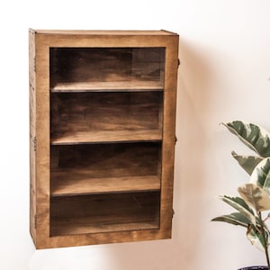 May include: A wooden wall-mounted shelf with three glass shelves and a glass door. The shelf is stained a dark brown color.