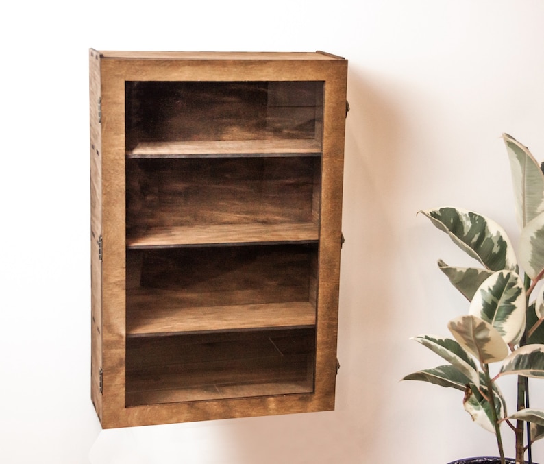 May include: A wooden wall-mounted shelf with three glass shelves and a glass door. The shelf is stained a dark brown color.