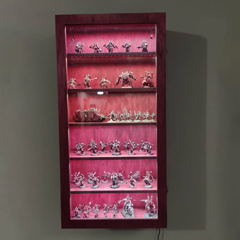 May include: A dark wood display case with glass shelves, lit from within, showcasing a collection of miniature figures. The figures are arranged in rows on each shelf, with a variety of poses and weapons. The case is a great way to display and protect a collection of miniature figures.