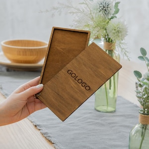 May include: A wooden box with a hinged lid, engraved with the word "GOLOGO". The box is open, showing the inside. The wood has a warm, brown colour. The background includes a table setting with a bowl and flowers.