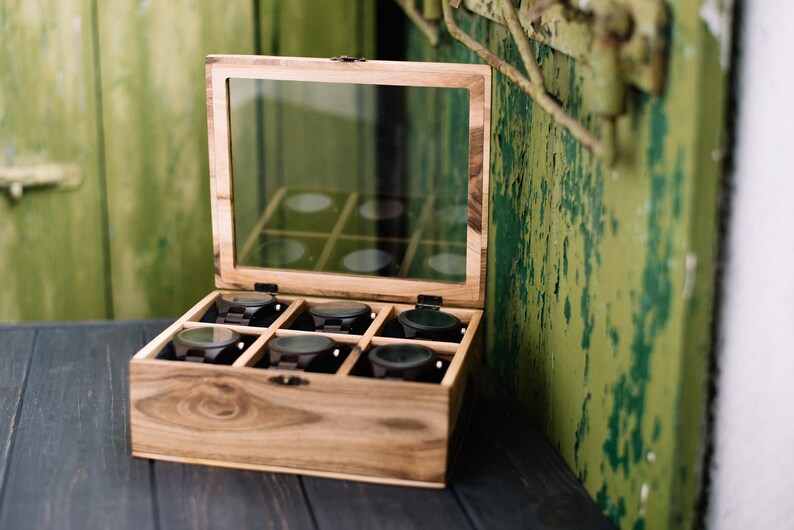 May include: A wooden watch box with a glass lid, open to reveal eight compartments, each containing a black wristwatch. The box is made of light brown wood with dark brown accents.