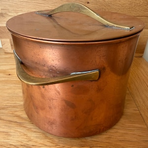 May include: A cylindrical copper pot with a matching lid and two curved brass handles. The exterior surface of the pot and lid shows a warm, metallic copper finish with subtle signs of wear and natural patina, while the brass handles are smooth.