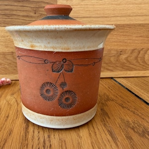 May include: A cylindrical terracotta ceramic canister with a matching lid. The body features a reddish-brown base with a cream-coloured glaze at the top and bottom rims. A black etched design depicts two circular floral patterns and leaves.