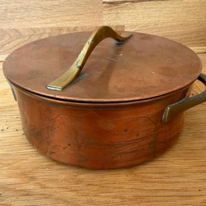 May include: A round copper pot with a matching lid, featuring a polished metallic finish and dark patina spots. The pot includes two curved brass handles attached to the sides, and the lid has a single arched brass handle on top.