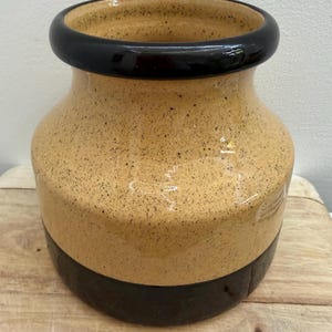 May include: A ceramic vase featuring a two-tone design with a glossy black rim and base, contrasted by a speckled tan middle section. The vessel has a wide, rounded body and a thick, smooth opening, showcasing a distinct colour-blocked aesthetic.