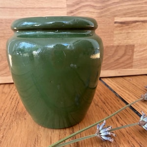 May include: A small, glossy, olive green ceramic jar with a rounded body and a thick, slightly protruding rim. The surface of the jar is smooth and reflective, showing subtle variations in the green glaze across its curved, bulbous shape.