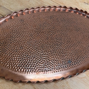 May include: An oval-shaped copper tray featuring a hammered texture across its surface. The tray has a scalloped edge with a raised rim, showcasing a metallic reddish-brown finish with subtle variations in colour throughout the metal surface.