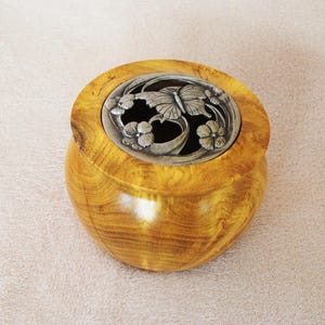 May include: A wooden container with a silver lid featuring a butterfly and floral design. The container has a rounded shape and a warm, golden-brown colour. The lid is intricately detailed with a butterfly and flowers.