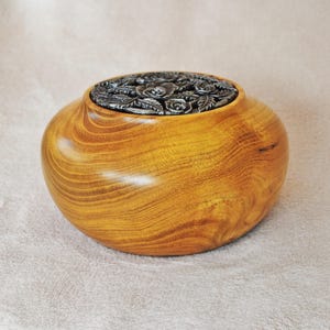 May include: A wooden bowl with a natural wood grain finish. The bowl has a dark metal lid with a floral rose design. The bowl is round and has a wide opening. The wood is a warm, golden colour.