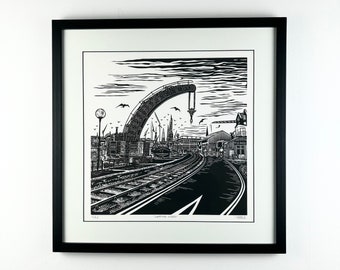 May include: Black and white woodcut print of a crane lifting a large object over a train track and a road. The print is framed in a black frame and has the text "Wapping Wharf" at the bottom.