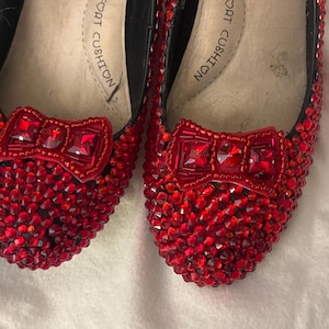 Könnte beinhalten: Ein Paar rote flache Schuhe, die vollständig mit kleinen roten Kristallen bedeckt sind. Jeder Schuh hat eine auffällige rote Schleife auf der Vorderkappe, die ebenfalls mit roten Kristallen verziert ist. Die hellen Innensohlen tragen den Aufdruck Comfort Cushion.