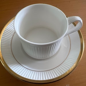 Może przedstawiać: Biały zestaw filiżanki i spodka ze złotą obwódką. Filiżanka ma żebrowany wzór, a spodek pasujący. Zestaw jest wykonany z porcelany i znajduje się na drewnianej powierzchni.