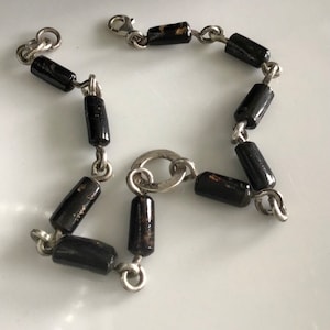 以下が含まれることがあります： 黒い円筒形のビーズが付いたシルバーカラーのブレスレット。ビーズは小さなシルバーのリンクと留め金で接続されています。ブレスレットは白い表面に表示されています。ビーズは光沢のある仕上がりです。