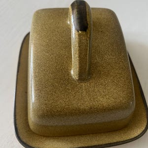 May include: A rectangular ceramic butter dish with a speckled olive-green glaze. The lid features a tall, arched handle with a dark brown accent at the top. The base has rounded corners and a dark brown rim, matching the handle's color tone.