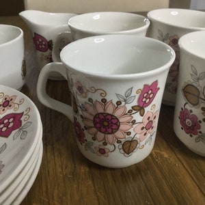 Puede incluir: Juego de té de cerámica blanca con un estampado floral en rosa, marrón y dorado. El juego incluye tazas, platillos y una jarra para crema. El diseño floral presenta grandes flores y hojas estilizadas.