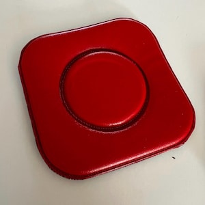 Pode incluir: Um porta-copos quadrado de cor vermelha brilhante com cantos arredondados e uma superfície lisa ligeiramente almofadada. O porta-copos apresenta uma secção central circular em relevo definida por uma borda costurada, criando um aspeto elevado.