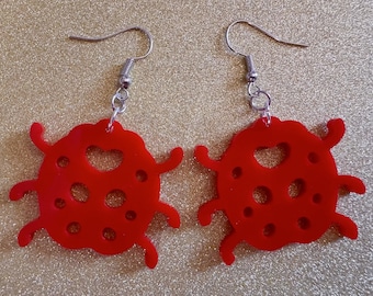 May include: A pair of red ladybug earrings with silver hooks. The ladybugs have a heart-shaped cutout and a dotted pattern.