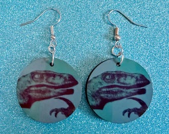 May include: A pair of round earrings featuring a dinosaur head design. The earrings have silver-colored hooks and are set against a sparkly blue background. The dinosaur image is a mix of red, purple, and blue.
