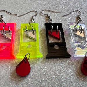 Puede incluir: Cuatro pendientes originales con forma de guillotinas, cada uno de un color diferente: rosa neón, amarillo neón, negro y transparente iridiscente. Cada pendiente tiene una cuchilla plateada y un dije en forma de lágrima.
